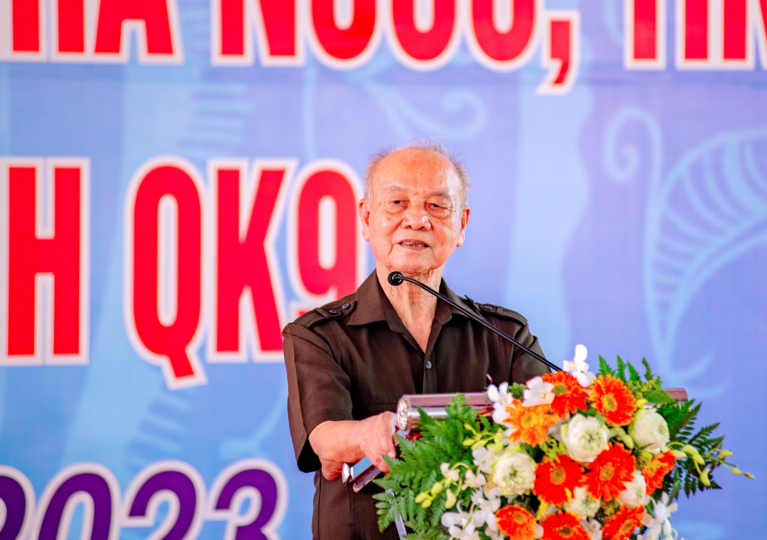 A person standing at a podium with a microphone and flowersDescription automatically generated